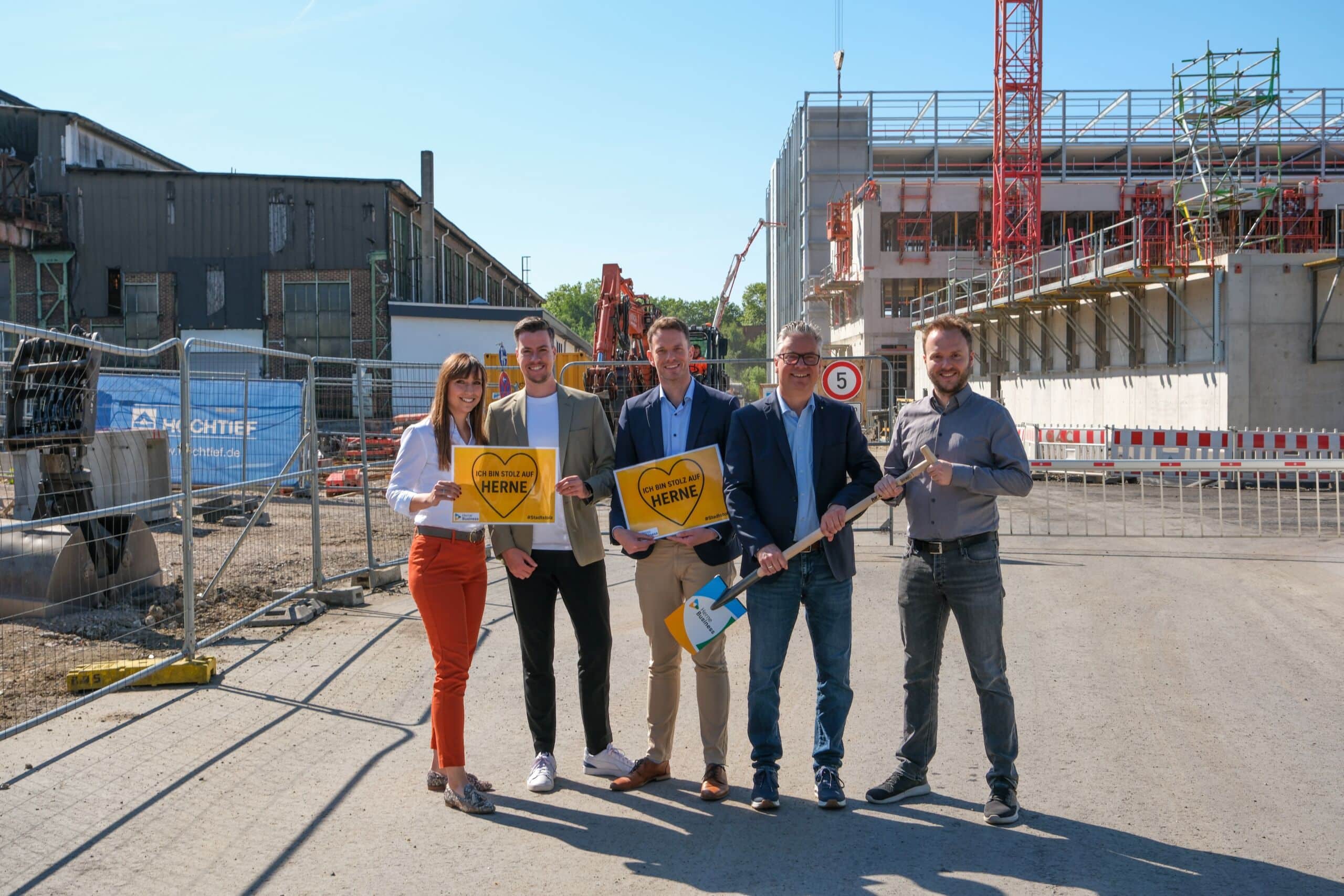 250704_Gruppenfoto FBQ Ost 2 Fünf Personen stehen im FunkenbergQuartier vor der Baustelle. Links ist das Bestandsgebäude des FunkenbergQuartier Ost zu sehen, rechts der Baufortschritt für die HSPV im FunkenbergQuartier West. Im Vordergrund stehen Dr. Svenja Rebsch, Lukas Issinger, Maurice Schirmer, Dr. Dirk Drenk und Fabian Schlosser. In der Hand halten sie zwei Schilder mit der Aufschrift „Ich bin stolz auf Herne“ sowie eine Schaufel mit der Aufschrift „Herne.Business“.