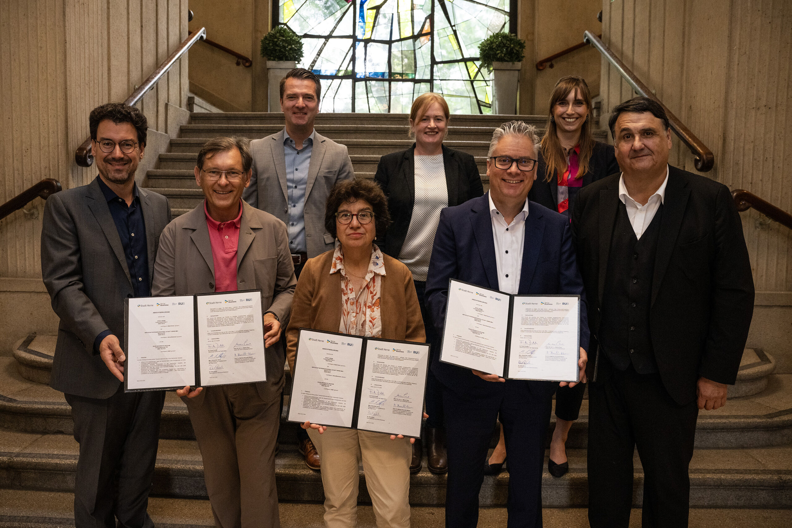 Personen stehen auf einer Treppe im Rathaus und präsentieren die unterzeichnete Absichtserklärung. Zu sehen sind im Vordergrund Marc Alexander Ulrich, Dr. Frank Dudda, Prof. Dr. Martina Havenith-Newen, Dr. Dirk Drenk und Prof. Dr. Dr. h. c. Martin Paul. Im Hintergrund sind Marc Seelbach, Dr. Rachel Glaves und Dr. Svenja Rebsch zu sehen.